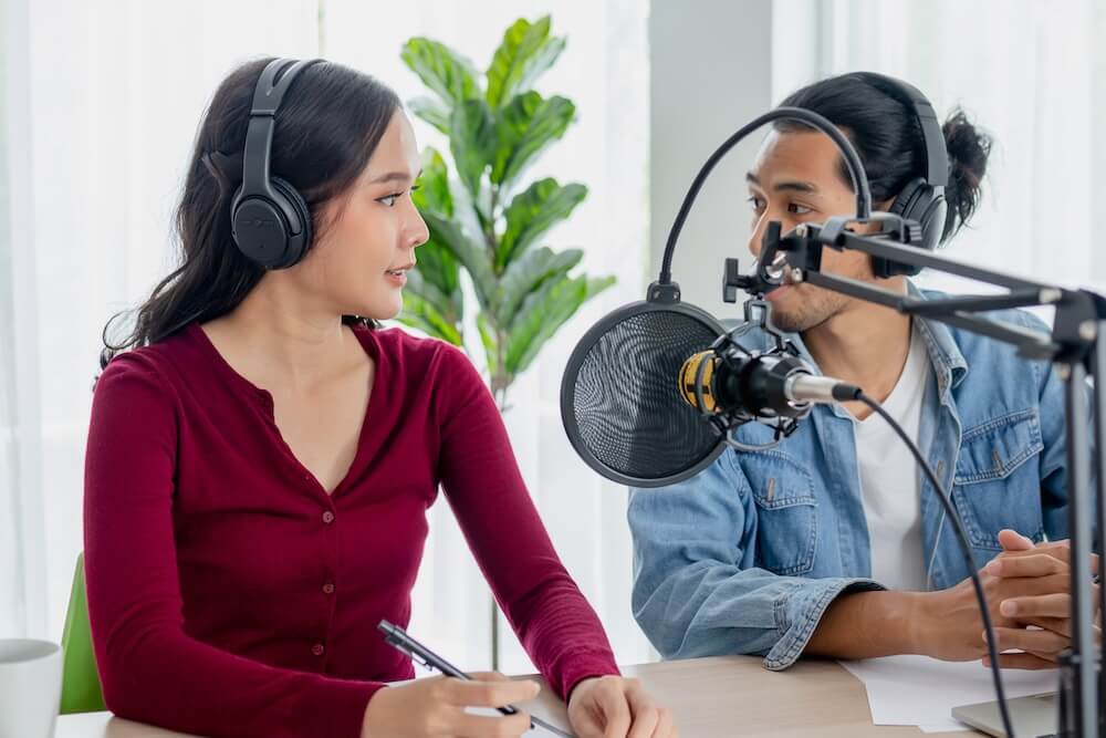 Two podcasters sit behind microphones in a white room, engaged in a discussion.