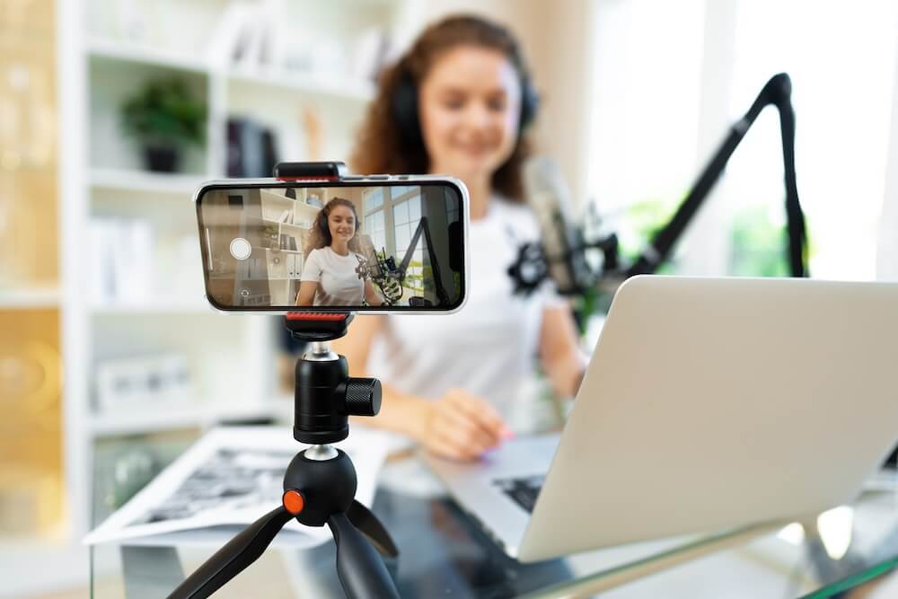 A person with curly hair records a podcast while filming themself on a smartphone with a small tripod.