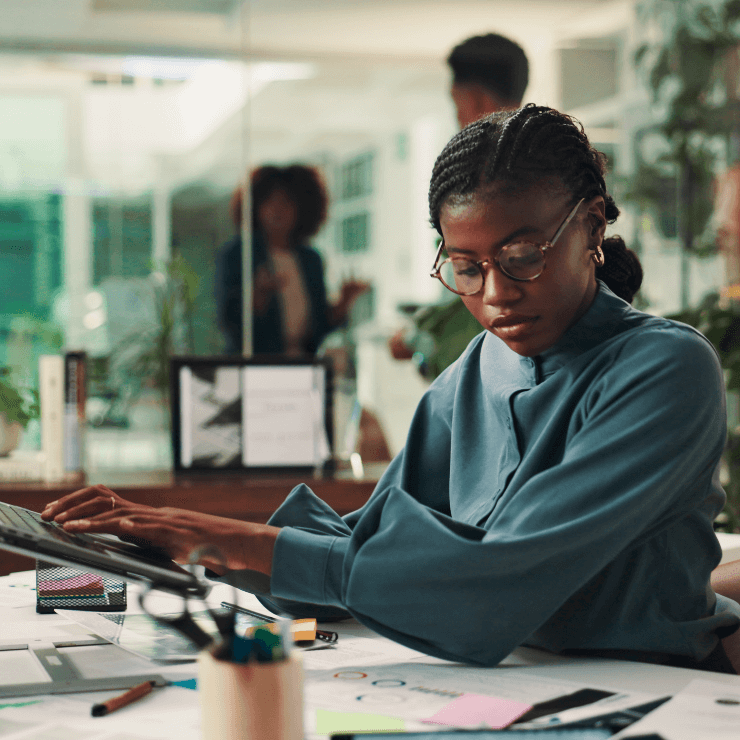 A person with glasses and braided hair sits at a desk covered with papers and uses a laptop in a modern office environment with several people standing in the background.
