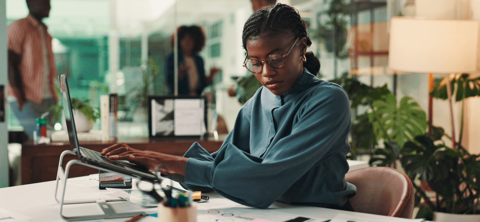 A person with glasses and braided hair sits at a desk covered with papers and uses a laptop in a modern office environment