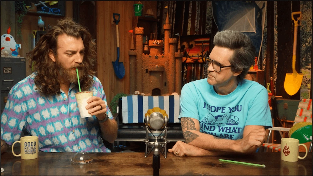 Screenshot of a YouTube video featuring Rhett and Link in a quirky, playhouse-style podcast studio while drinking smoothies.