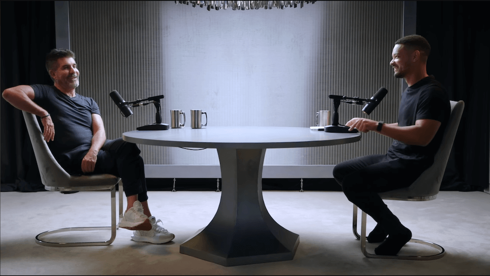 Screenshot of a YouTube video of Simon Cowell and Steven Bartlett sitting across a table from one another in a minimalist podcast studio.