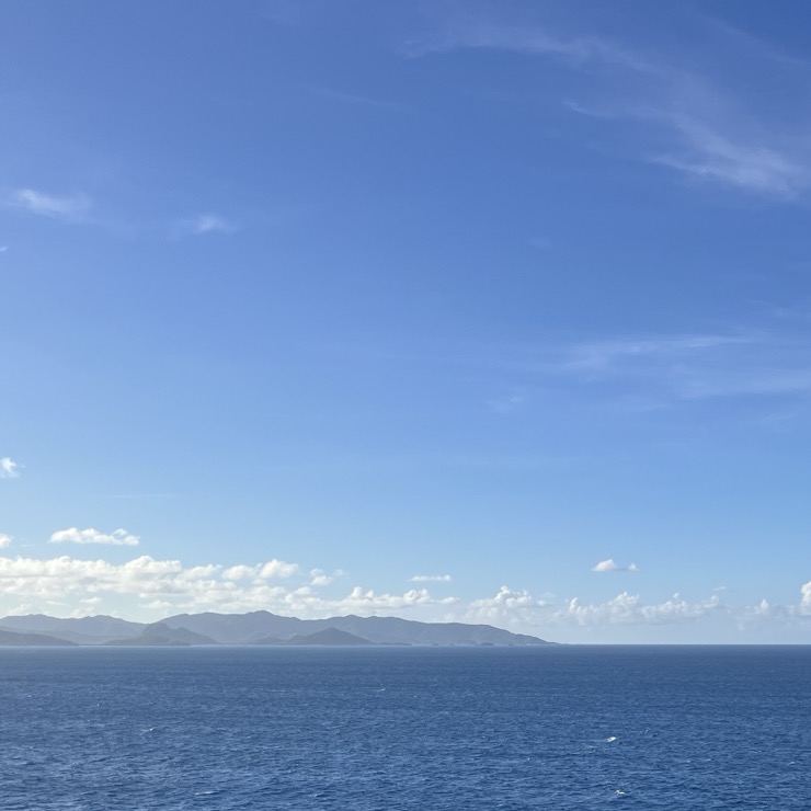 Blue Caribbean sea and huge blue sky with soft clouds. A mountainous island is on the horizon between sea and sky.
